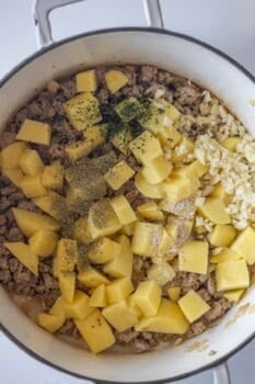 Cubed potatoes, ground meat, chopped garlic, and seasoning are shown in a large pot, ready to be cooked together for a flavorful pickle sausage soup.