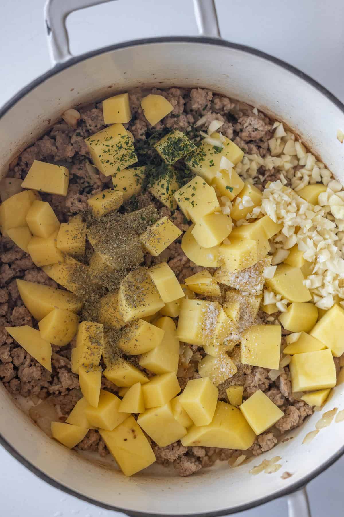 Cubed potatoes, ground meat, chopped garlic, and seasoning are shown in a large pot, ready to be cooked together for a flavorful pickle sausage soup.