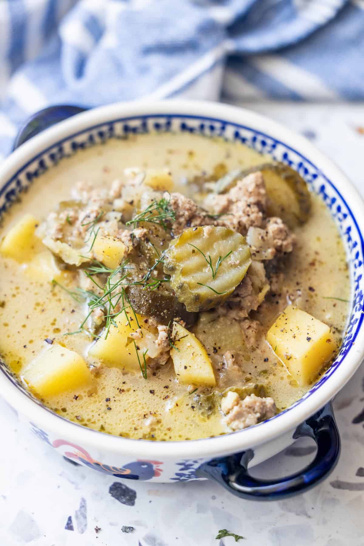 A bowl of creamy sausage soup containing ground meat, potato chunks, sliced pickles, and fresh herbs, served in a patterned bowl on a light surface.