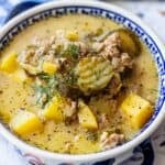 A bowl of creamy soup with ground meat, diced yellow potatoes, tangy pickles, and fresh dill is served in a decorative bowl, creating a comforting dish that combines classic soup flavors with a hint of pickle zest.