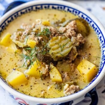 A bowl of creamy soup with ground meat, diced yellow potatoes, tangy pickles, and fresh dill is served in a decorative bowl, creating a comforting dish that combines classic soup flavors with a hint of pickle zest.