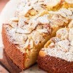 A slice of almond cake topped with sliced almonds and powdered sugar is lifted from the whole dessert on a metal server.