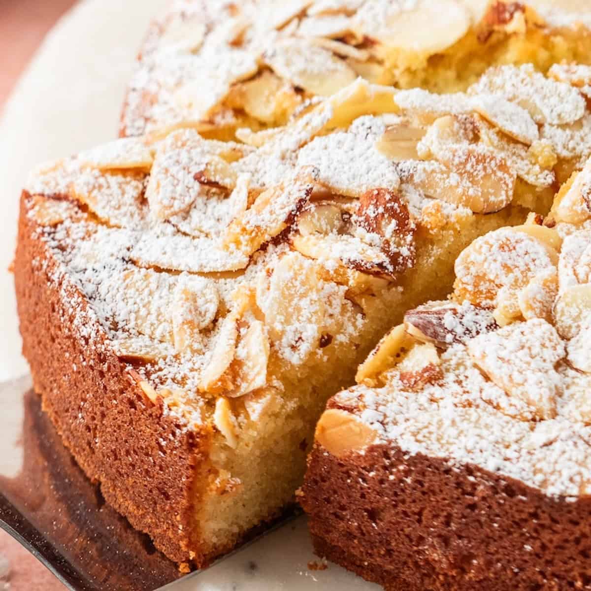 A slice of almond cake topped with sliced almonds and powdered sugar is lifted from the whole dessert on a metal server.