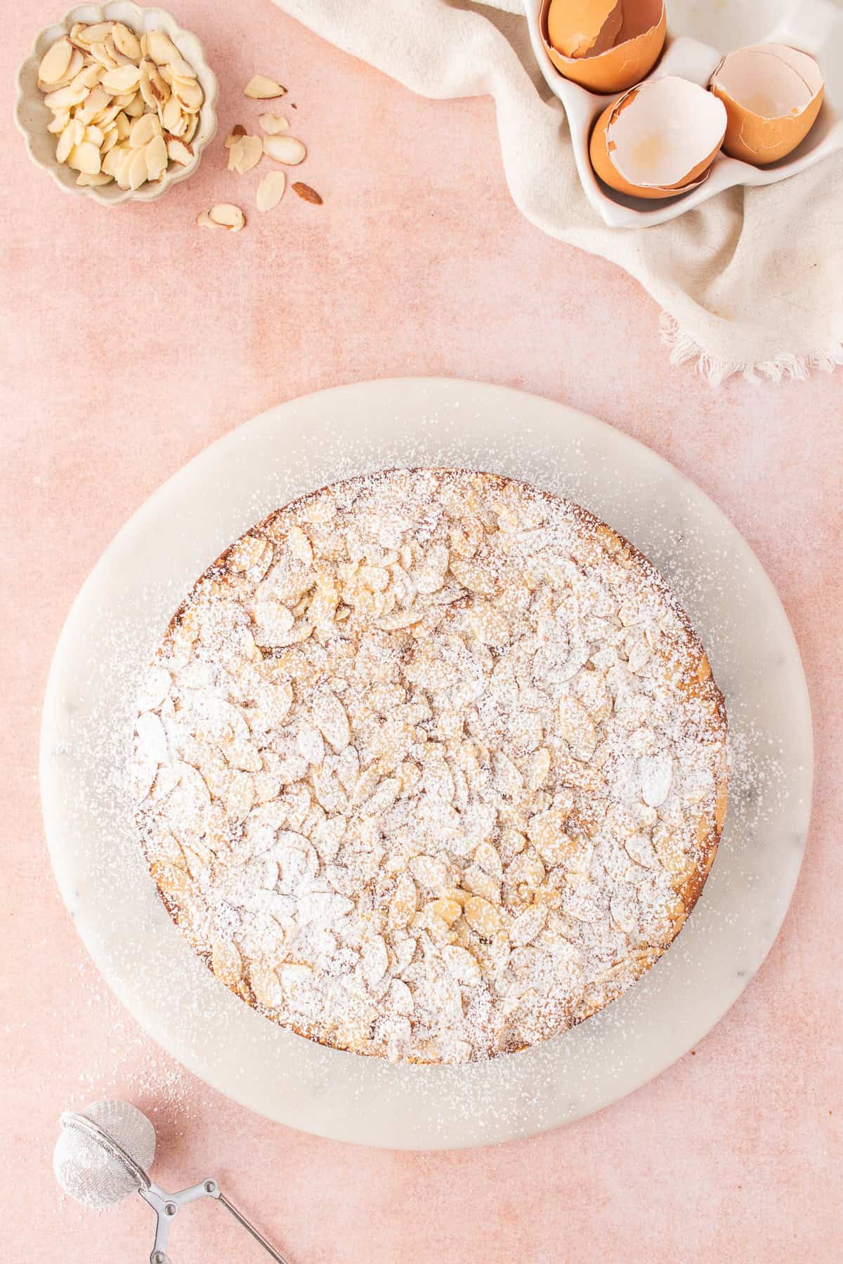 An easy almond cake topped with sliced almonds and powdered sugar on a marble board, with bowls of almond slivers, eggshells, and a sifter nearby.
