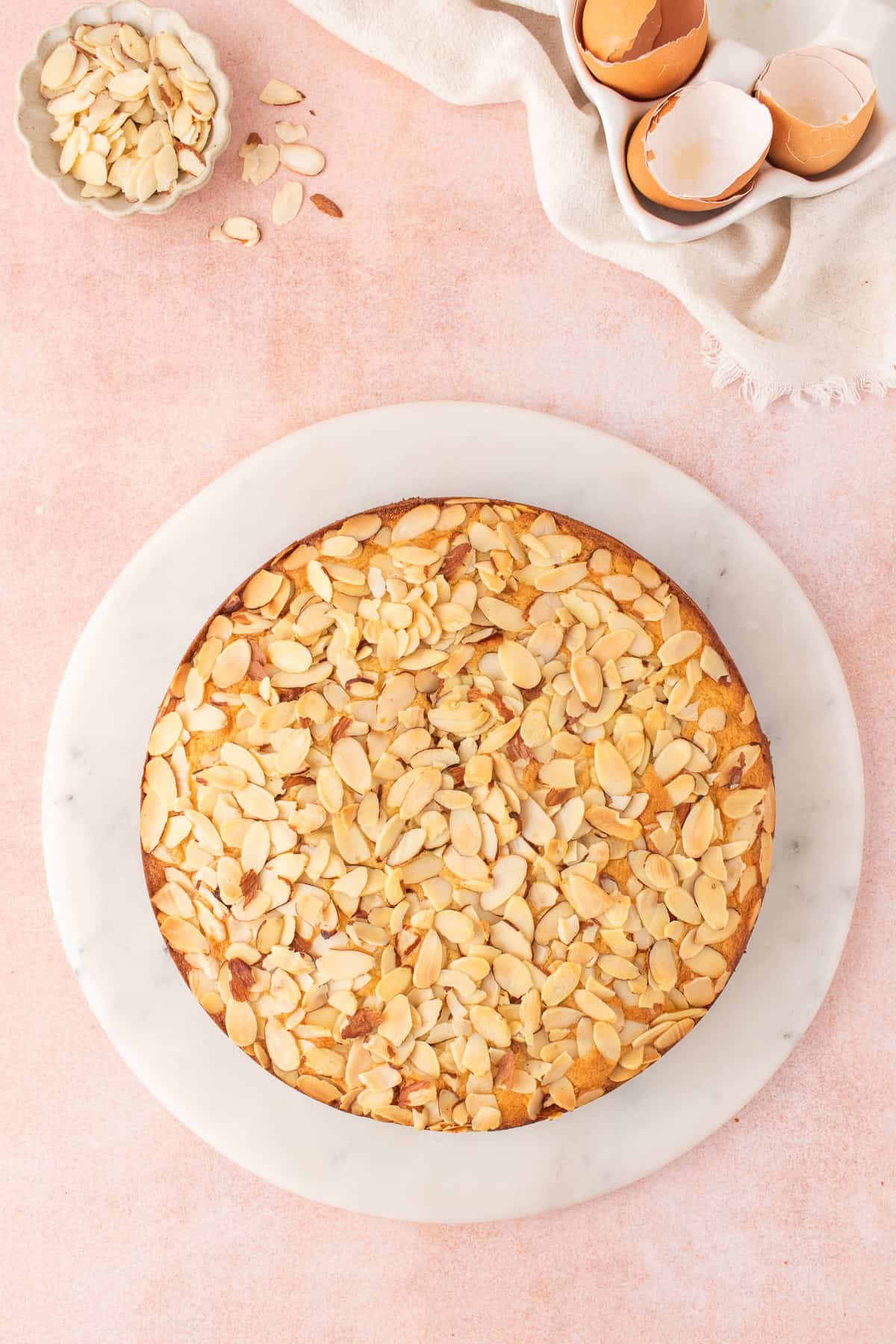 A round almond cake topped with sliced almonds on a marble serving board, with a small bowl of almonds and eggshells nearby.