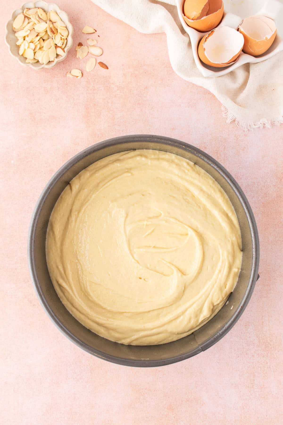 Cake batter in a round metal baking pan for an easy almond cake, with a small bowl of sliced almonds and cracked eggshells nearby.