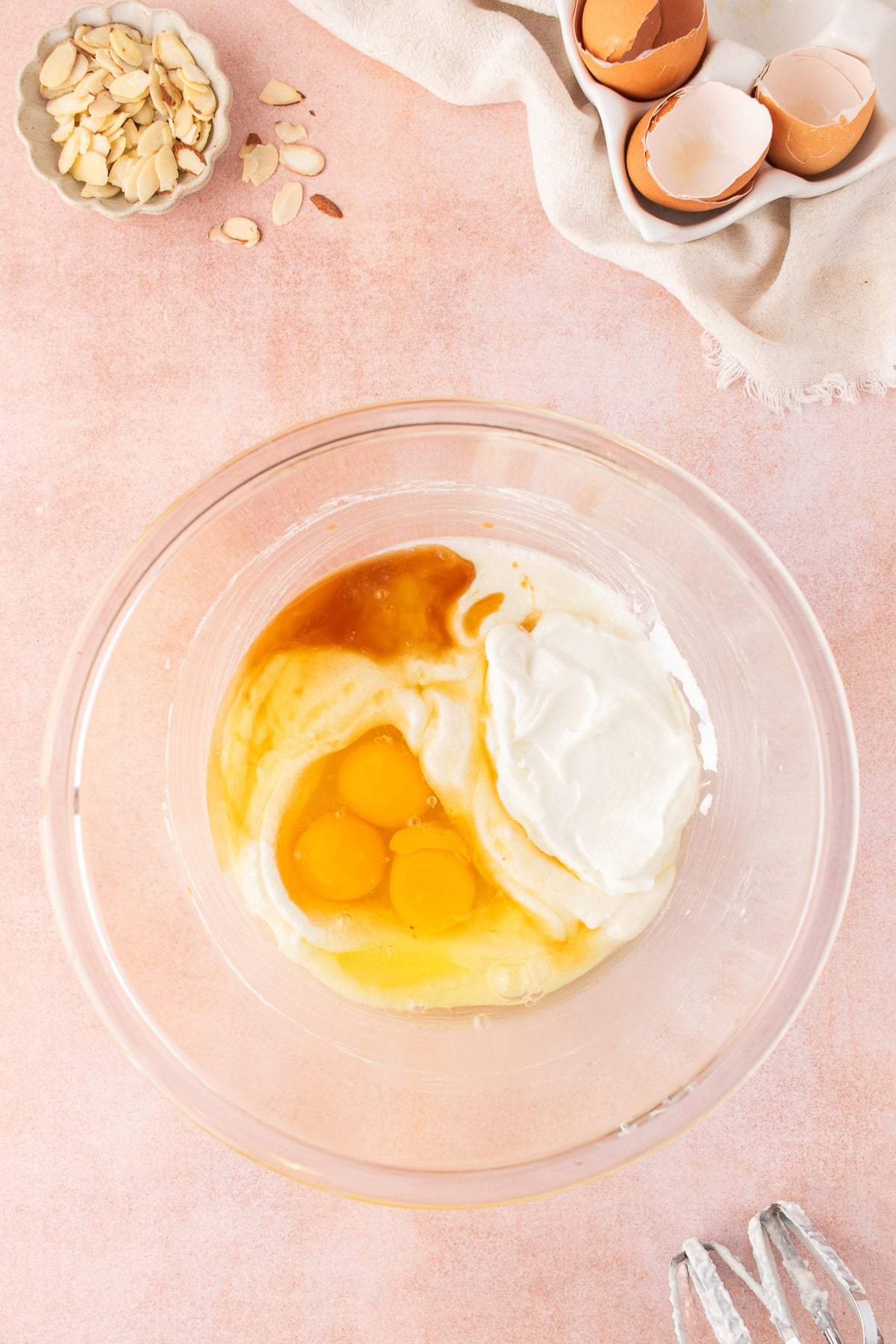 Glass bowl containing ingredients for an almond cake, including eggs, yogurt, and vanilla extract, with a small bowl of sliced almonds and eggshells nearby.