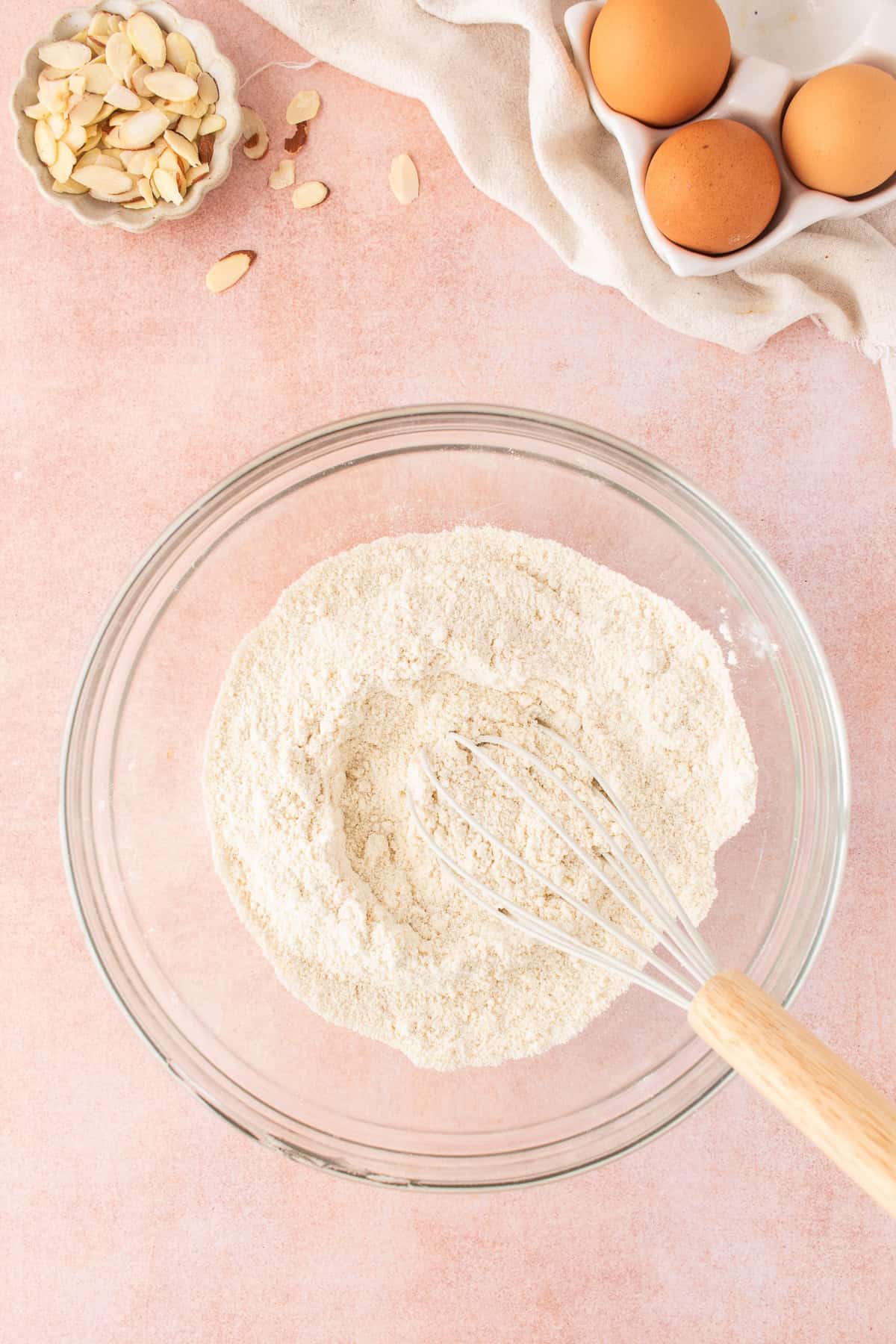 A glass bowl containing flour and a whisk, with a small bowl of sliced almonds and a carton of eggs nearby on a pink surface for an easy almond cake recipe.