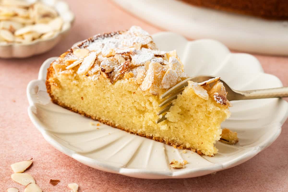 A slice of almond cake topped with sliced almonds and powdered sugar on a scalloped plate, with a fork breaking off a piece of the easy almond cake.