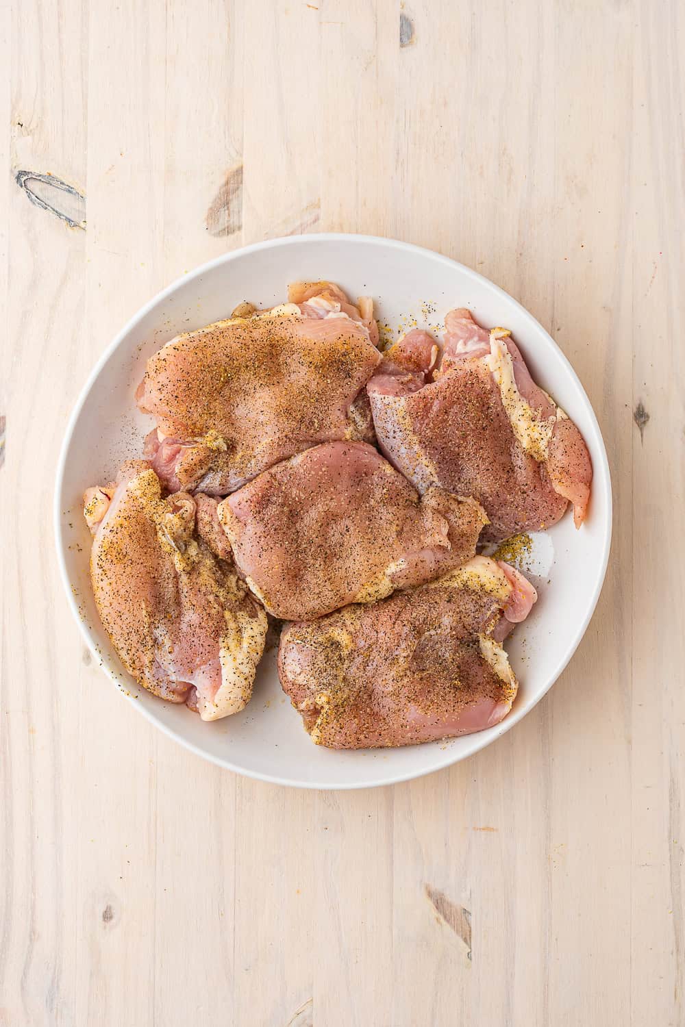 Four raw, seasoned chicken thighs in a white bowl on a wooden surface, ready for a one pot chicken and rice recipe.