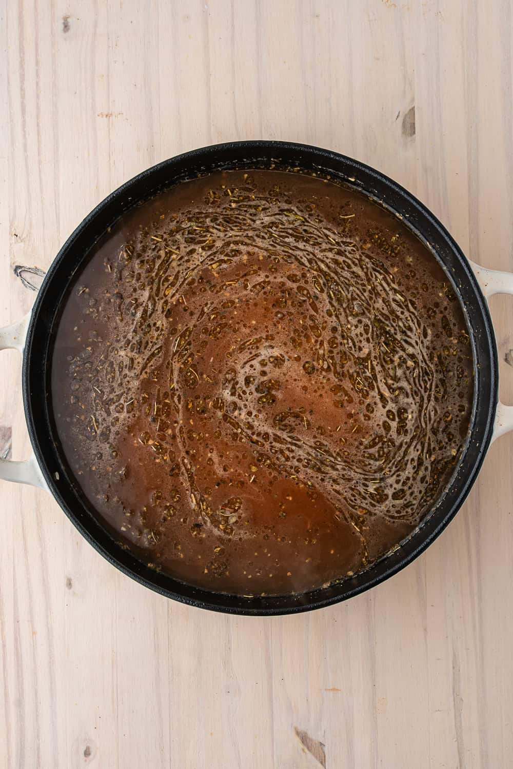 A shallow pan containing a dark, simmering liquid with visible herbs and oils for a one pot chicken dish, resting on a wooden surface.