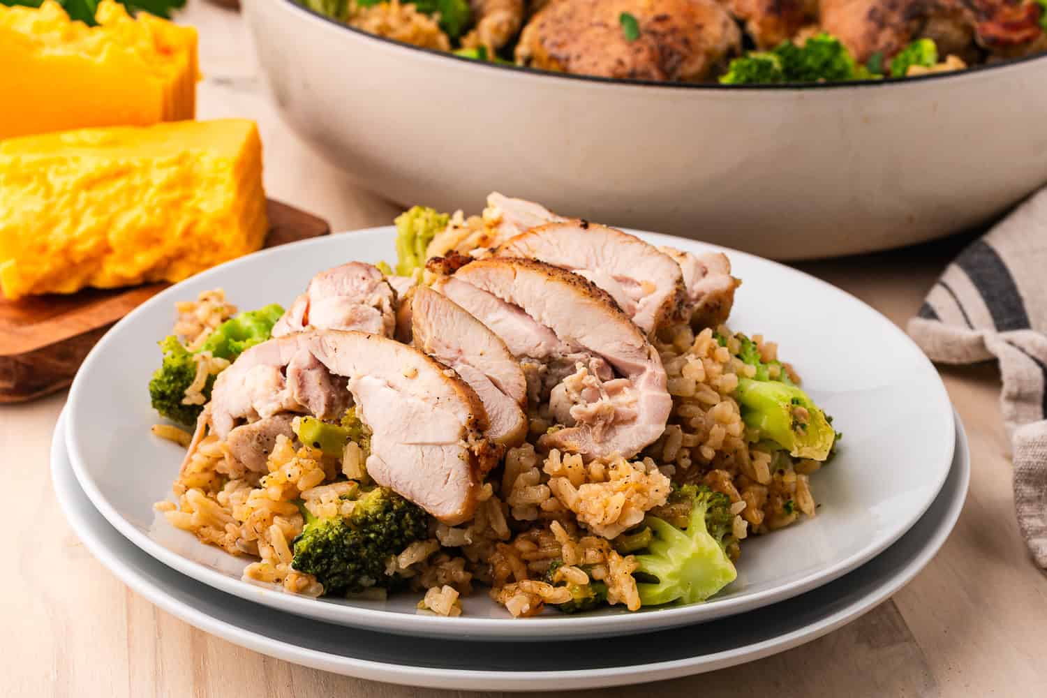 Sliced chicken breast served over brown rice and broccoli, with a pan of cheesy broccoli chicken and cheese blocks in the background.