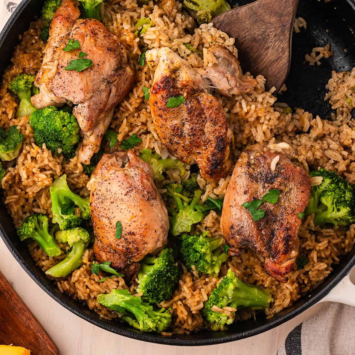 A skillet of cheesy chicken broccoli with brown rice and four seasoned chicken thighs, with a wooden spatula resting in the pan.