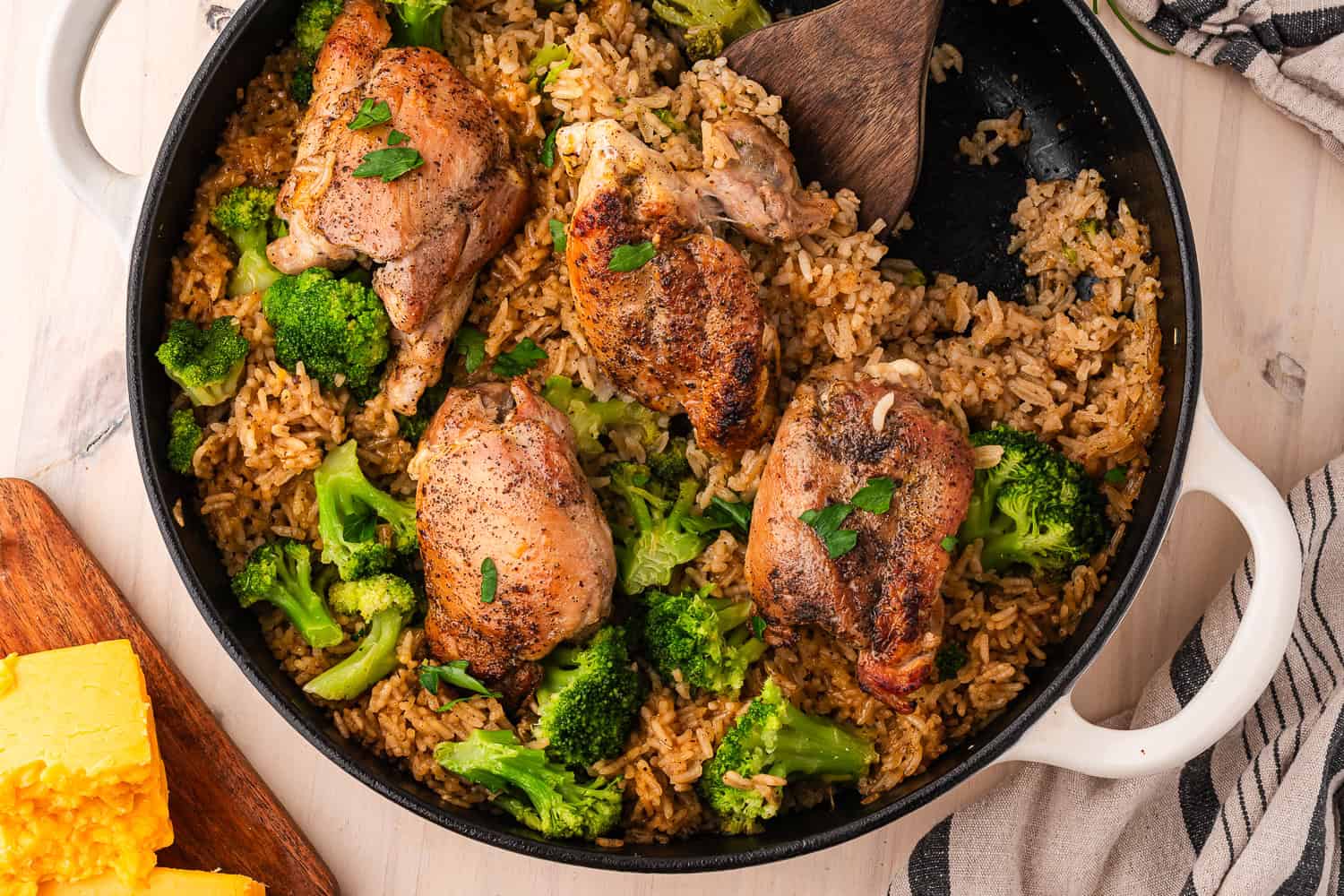 A skillet of cheesy chicken and broccoli with seasoned rice, featuring a wooden spatula and a side of cornbread.