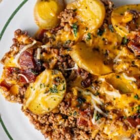 A plate of cheesy potato bake featuring sliced potatoes, ground meat, and bacon topped with melted cheese and herbs.