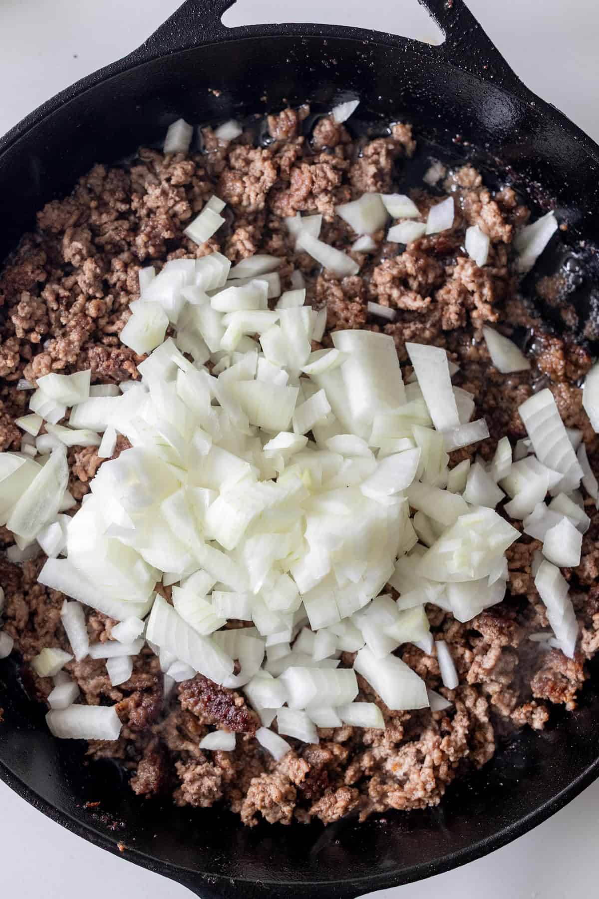 Ground beef browning in a cast iron skillet for a beef casserole, topped with a mound of chopped raw onion.