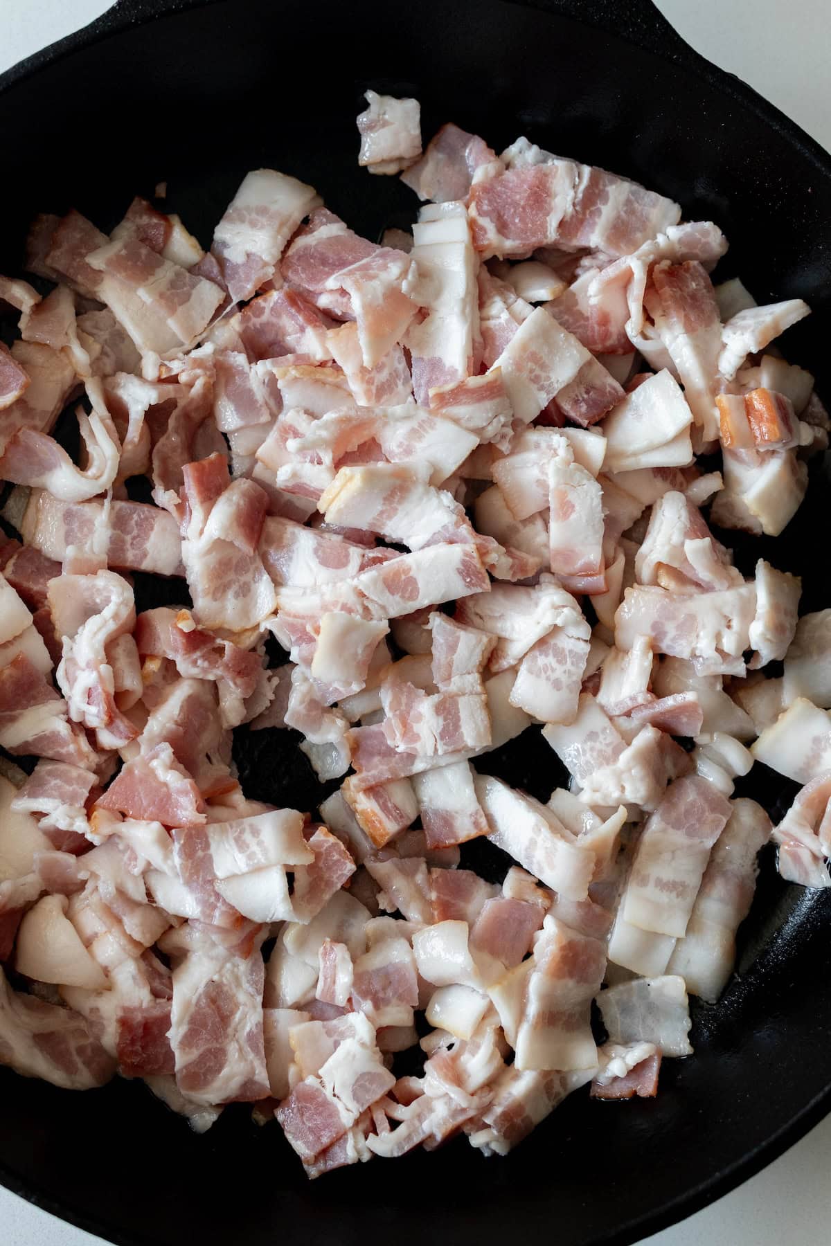 Chopped raw bacon pieces in a black cast-iron skillet, ready for a cheesy potato bake.