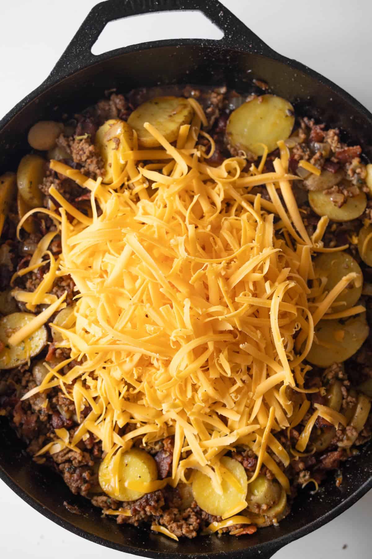 Cast iron skillet filled with a cheesy potato bake of ground meat, sliced potatoes, and shredded cheddar cheese.