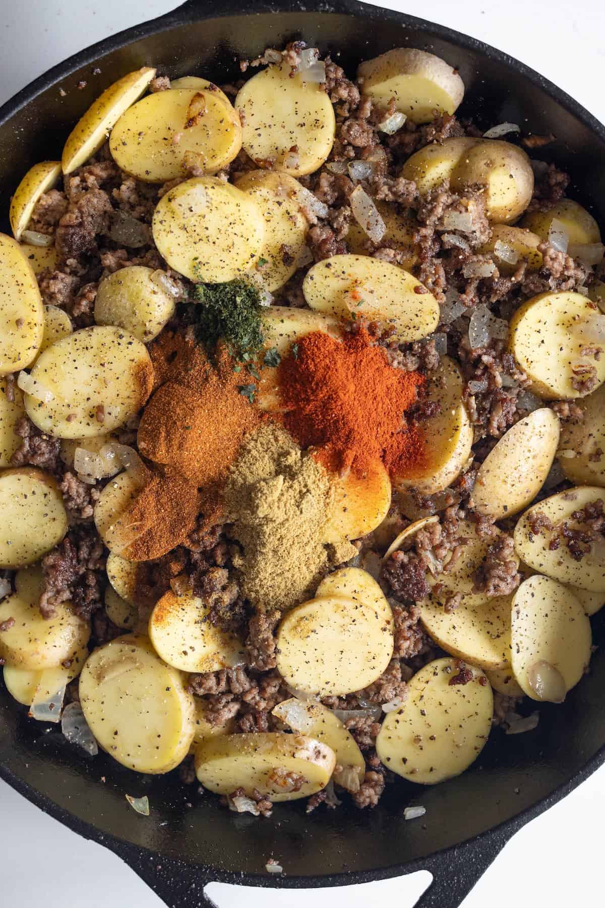 Sliced potatoes, ground meat, and onions in a cast iron skillet, forming a hearty potato and beef casserole with piles of ground spices and herbs on top.