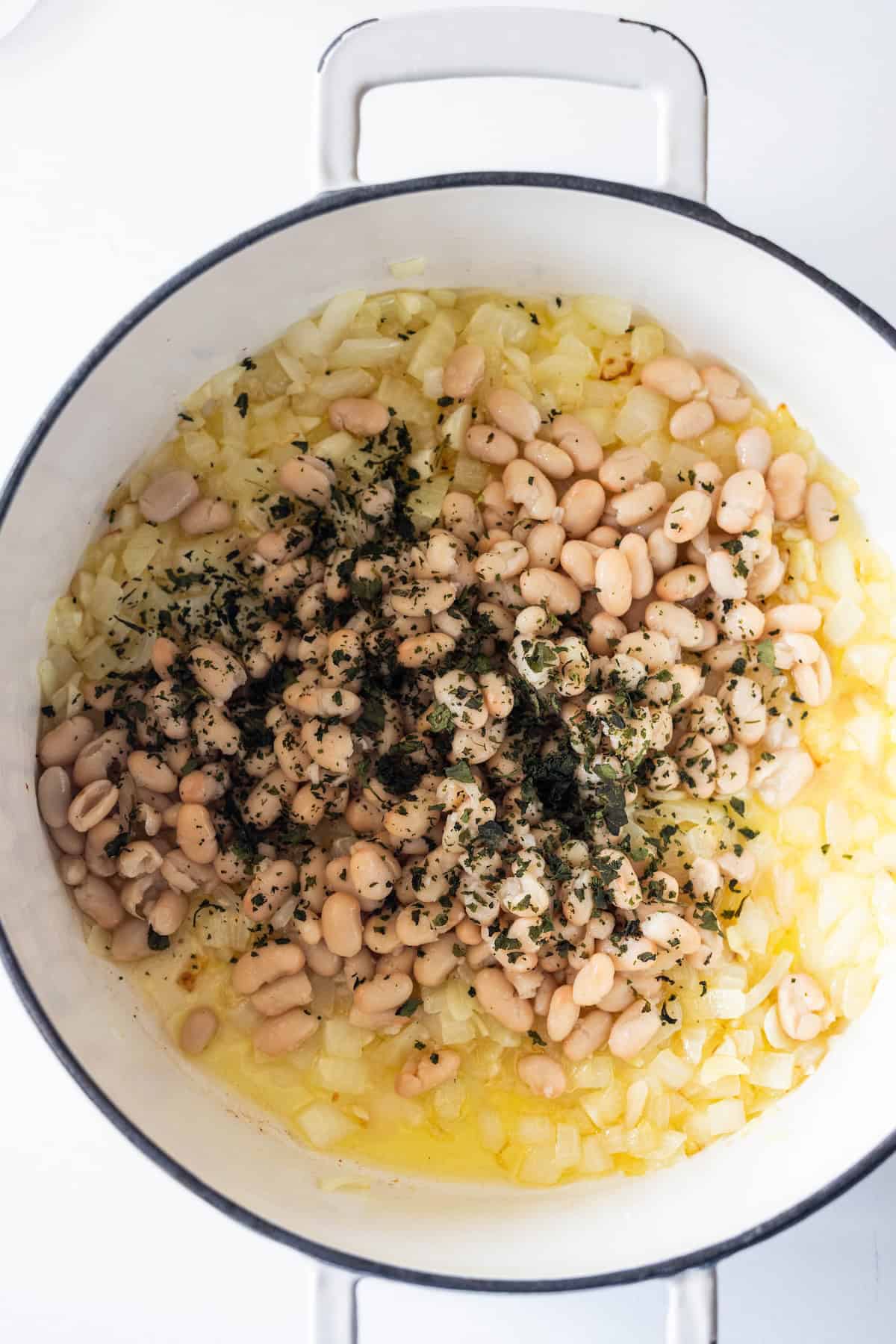 White cannellini beans, sautรฉed onions, and herbs simmered in a white enameled pot as part of a frozen vegetable soup.