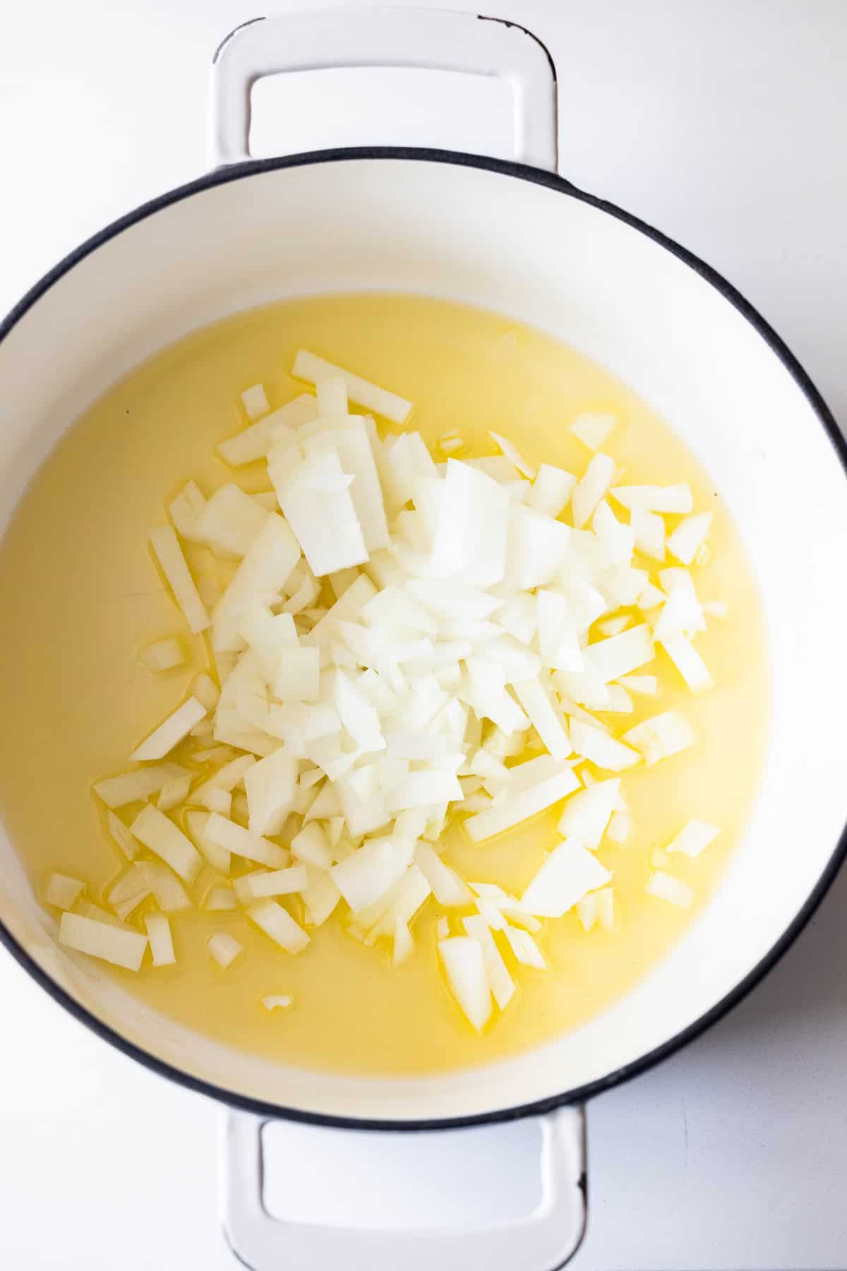 Diced white onions sautรฉing in oil at the bottom of a white pot for vegetable soup.