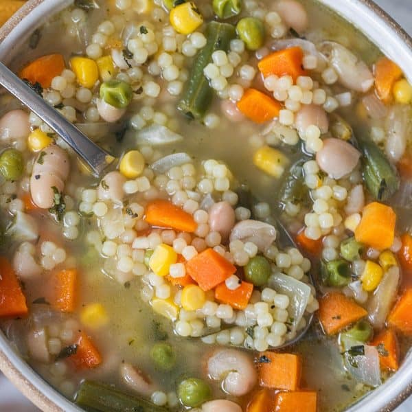 A bowl of frozen vegetable soup with pasta pearls, carrots, corn, peas, green beans, and white beans, with a spoon resting in the broth.