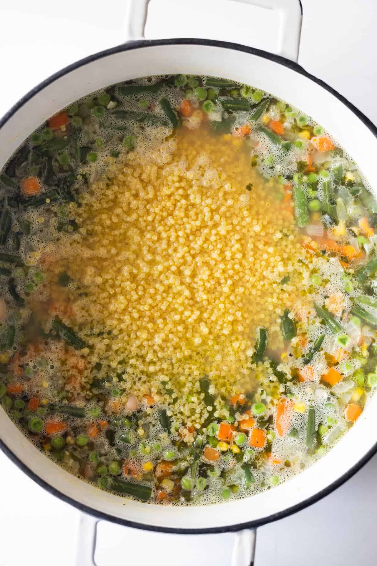A Dutch oven filled with frozen vegetable soup containing carrots, green beans, peas, and a cluster of small pasta in the center.