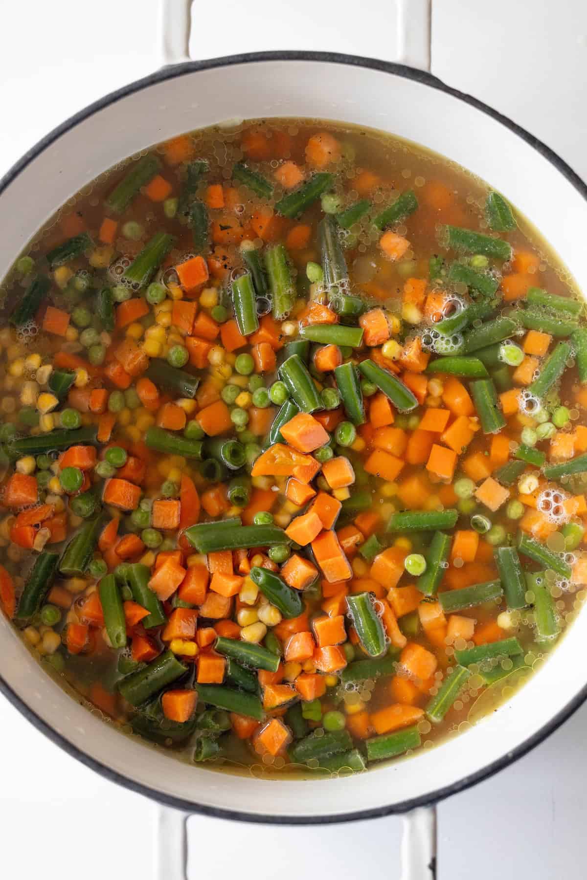A white pot filled with frozen vegetable soup containing diced carrots, green beans, peas, and corn in a clear broth.