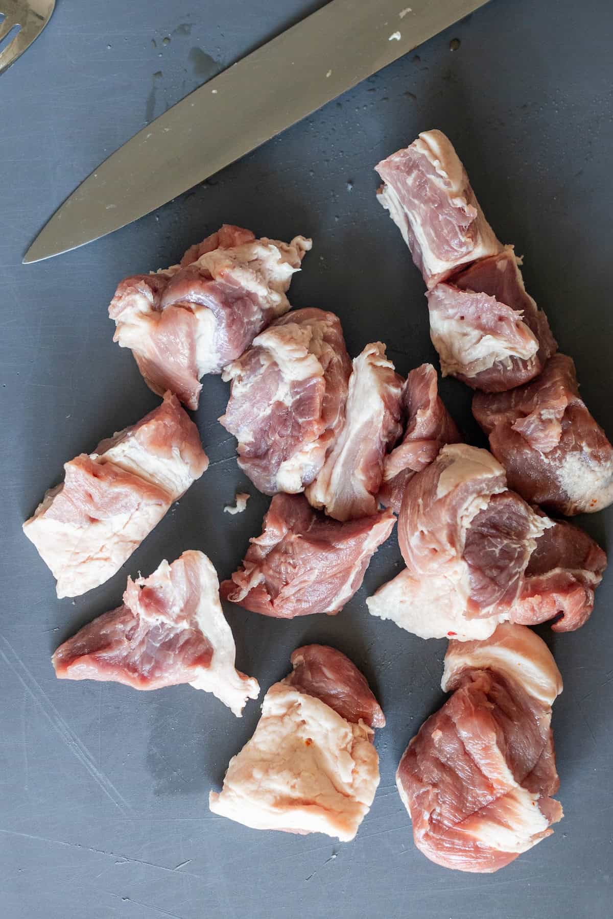 Chunks of onion marinated pork on a grey cutting board next to a chef’s knife.